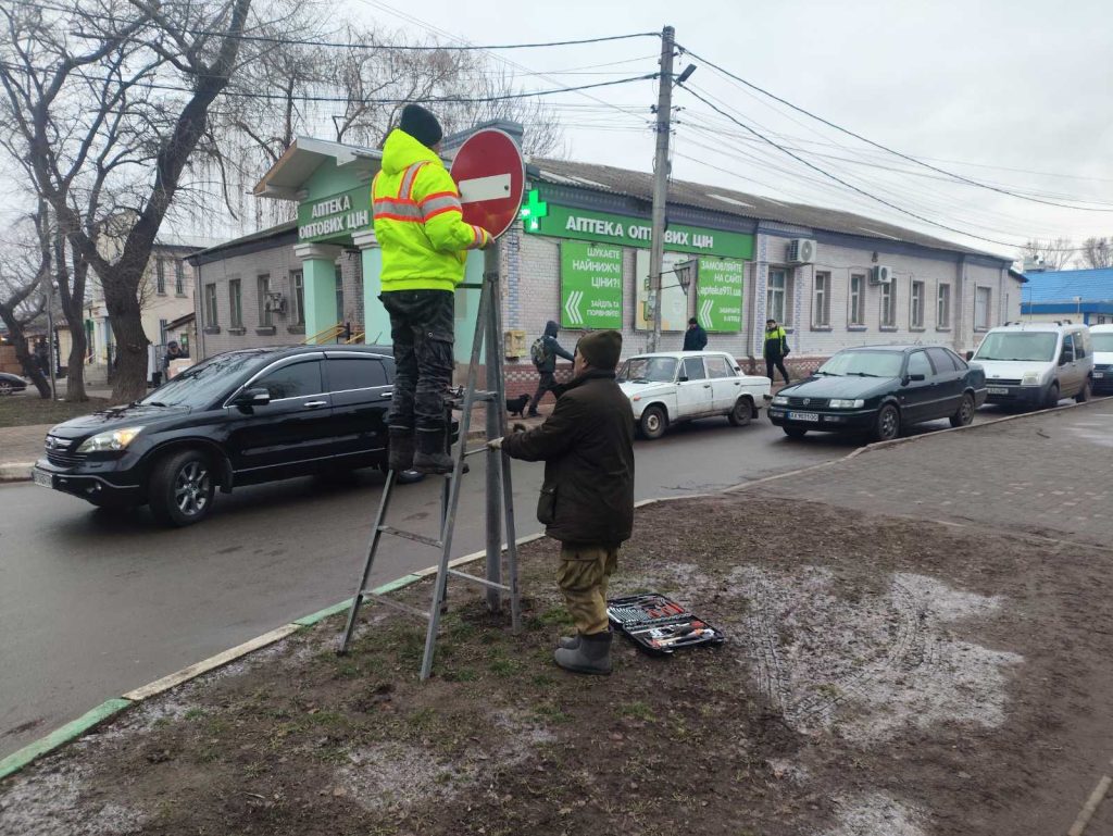 Два чоловіки, один на драбині, а інший біля неї, встановлюють червоний дорожній знак на стовпі обабіч дороги, де припарковані автомобілі, а на фоні видніється будівля аптеки.