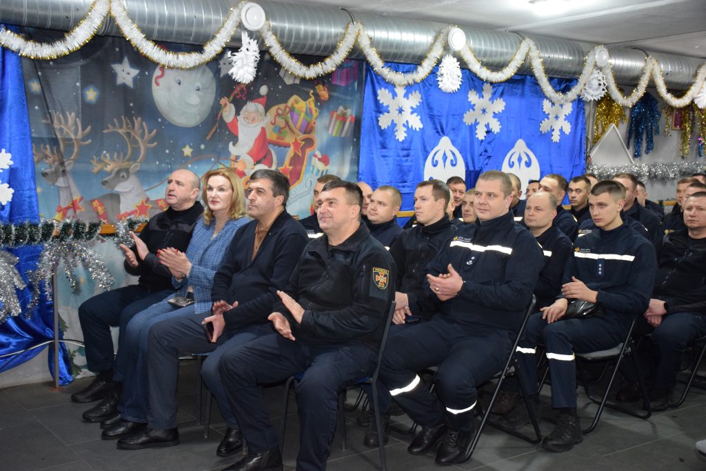 Група людей, серед яких чоловіки в уніформі та жінка, сидить і аплодує на тлі святкових новорічних декорацій з гірляндами, сніжинками та тематичними зображеннями.
