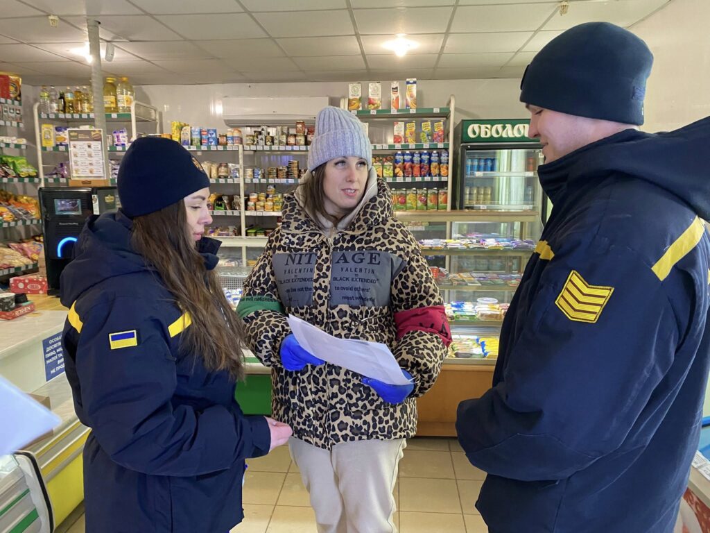 Жінка у світлому сірому біні та леопардовому пальто із чорними латками, імовірно, спілкується із двома військовими у формі із жовтими відзнаками.