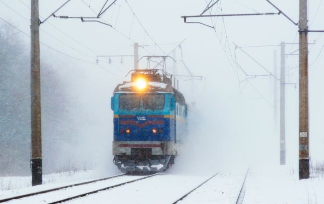 Блакитно жовтий потяг з яскравим прожектором рухається засніженою залізничною колією серед електричних стовпів під час снігопаду.