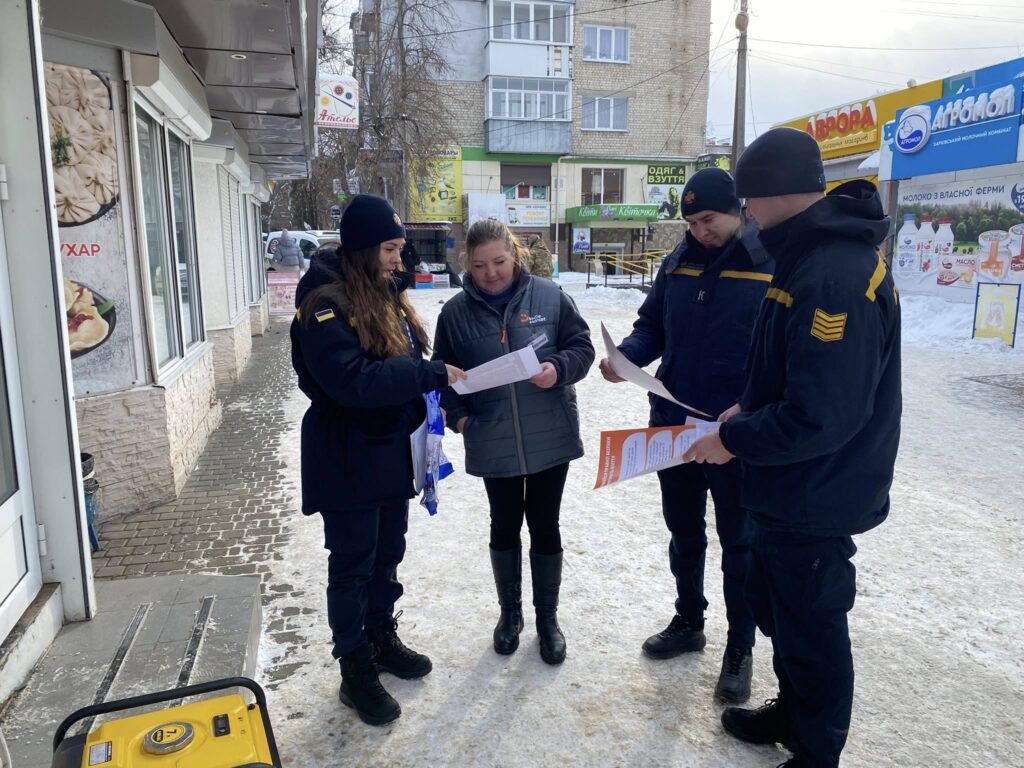 Жінка стоїть на снігу біля двох чоловіків у формі, розглядаючи папери, які вони тримають у руках.
