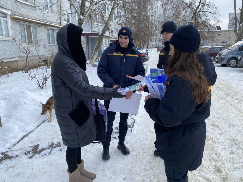 Жінка у довгій чорній пуховику отримує папери від жінки у чорній куртці та шапці, троє людей у чорно-жовтій формі спостерігають.
