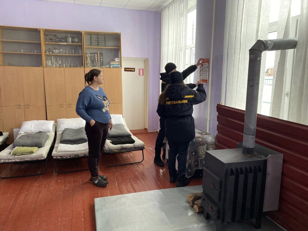 Жінка у формі рятувальника кріпить вогнегасник на стіні приміщення, де встановлена пічка та облаштовані місця для ночівлі.