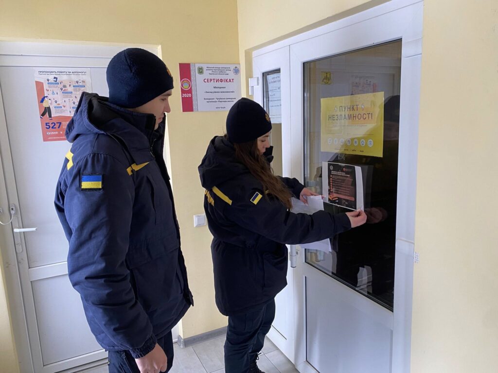 Жінка та чоловік у чорних хутряних куртках із жовтими та синьо-жовтими нашивками на рукавах наклеюють інформаційний плакат на скло білого дверного блоку із жовтим папером із написом