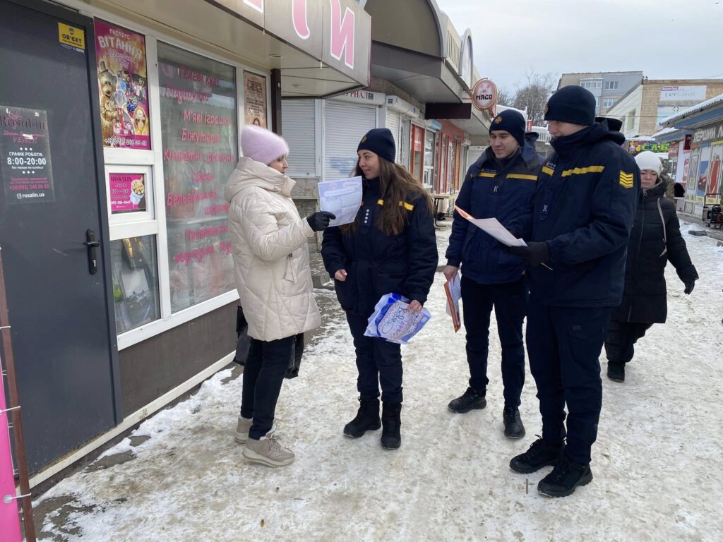 Жінка в білій шубі показує групі людей у чорній формі з жовтими смугами папери, що вони розглядають на снігу біля будівлі з інформаційними плакатами.