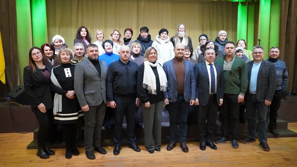 Велика група дорослих чоловіків і жінок стоїть рядами на деревяній підлозі перед сценою з зеленими та жовтими шторами, позуючи для спільного фото.