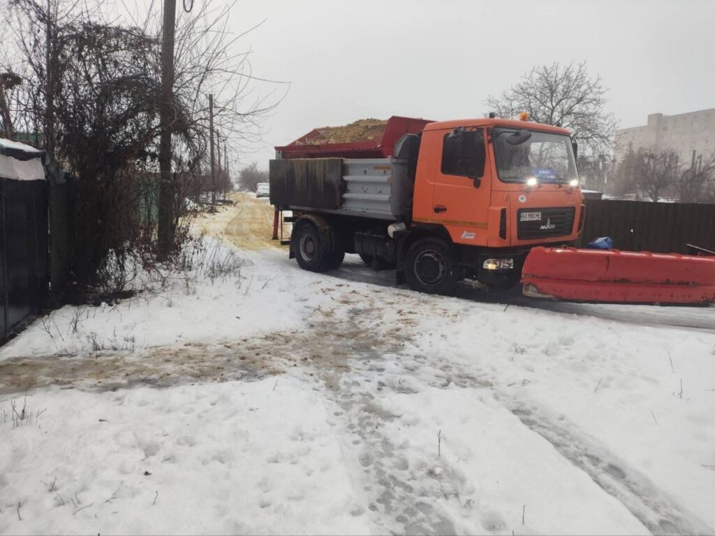 Помаранчевий снігоприбиральний автомобіль із відвалом та піднятим кузовом, заповненим піском, їде по сніжній дорозі.
