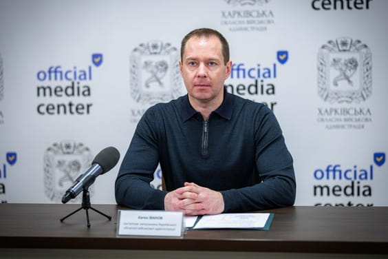 Чоловік сидить за столом із мікрофоном, складавши руки перед собою та дивлячись у камеру, навпроти нього табличка з його іменем та інший папка з документами.