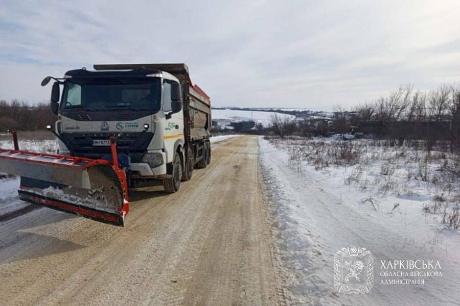 Велика біла вантажівка з снігоприбиральною машиною на переді їде по сільській дорозі вкритої снігом.