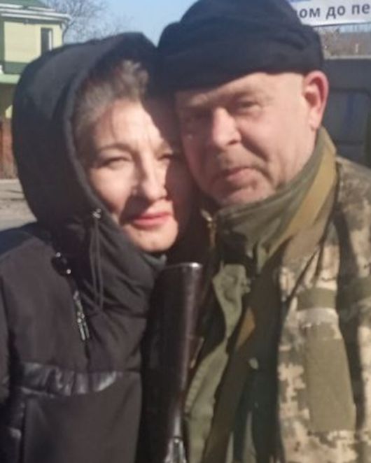 Жінка у чорній куртці зі світло-сірим волоссям обіймає чоловіка у військовій формі та чорній шапці.