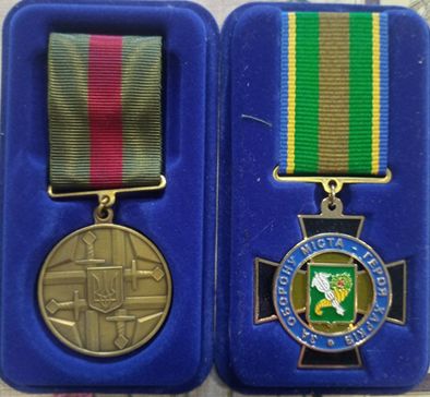 Дві медалі у відкритих синіх коробках із різнобарвними стрічками, на одній медалі зображено герб із хрестом та написом українською мовою.