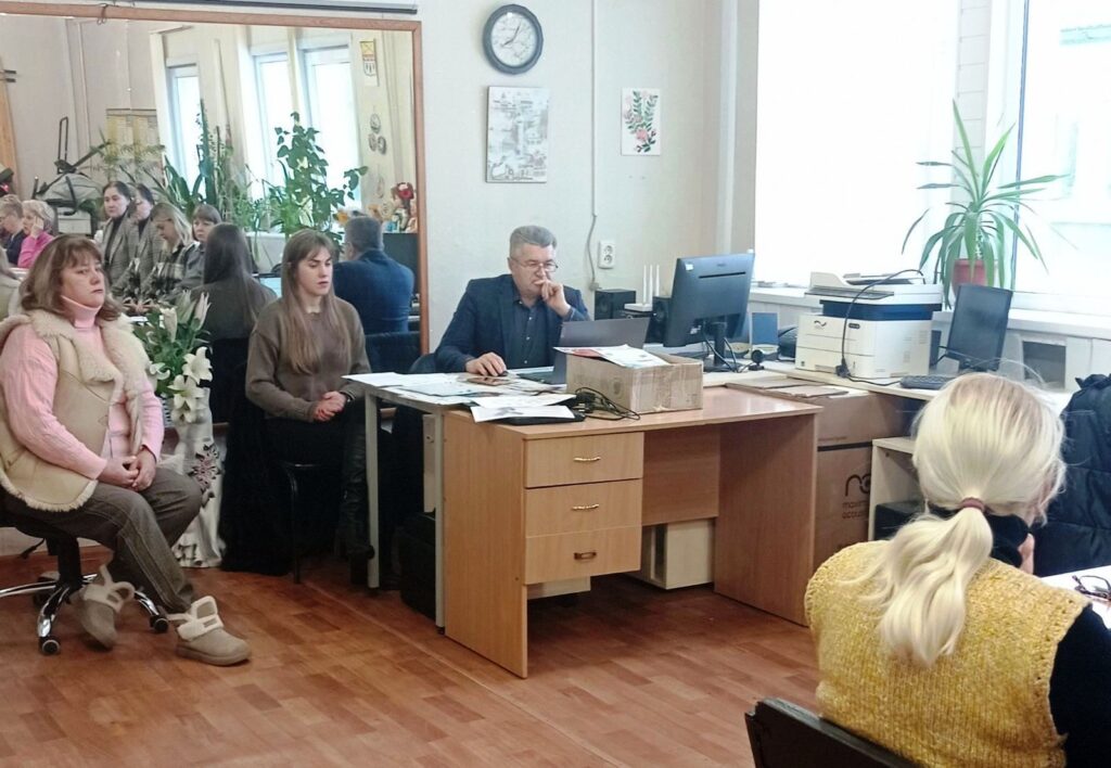 Чоловік сидить за столом і спирається рукою на підборіддя, поруч із ним дві жінки, ще одна жінка сидить спиною до глядача.
