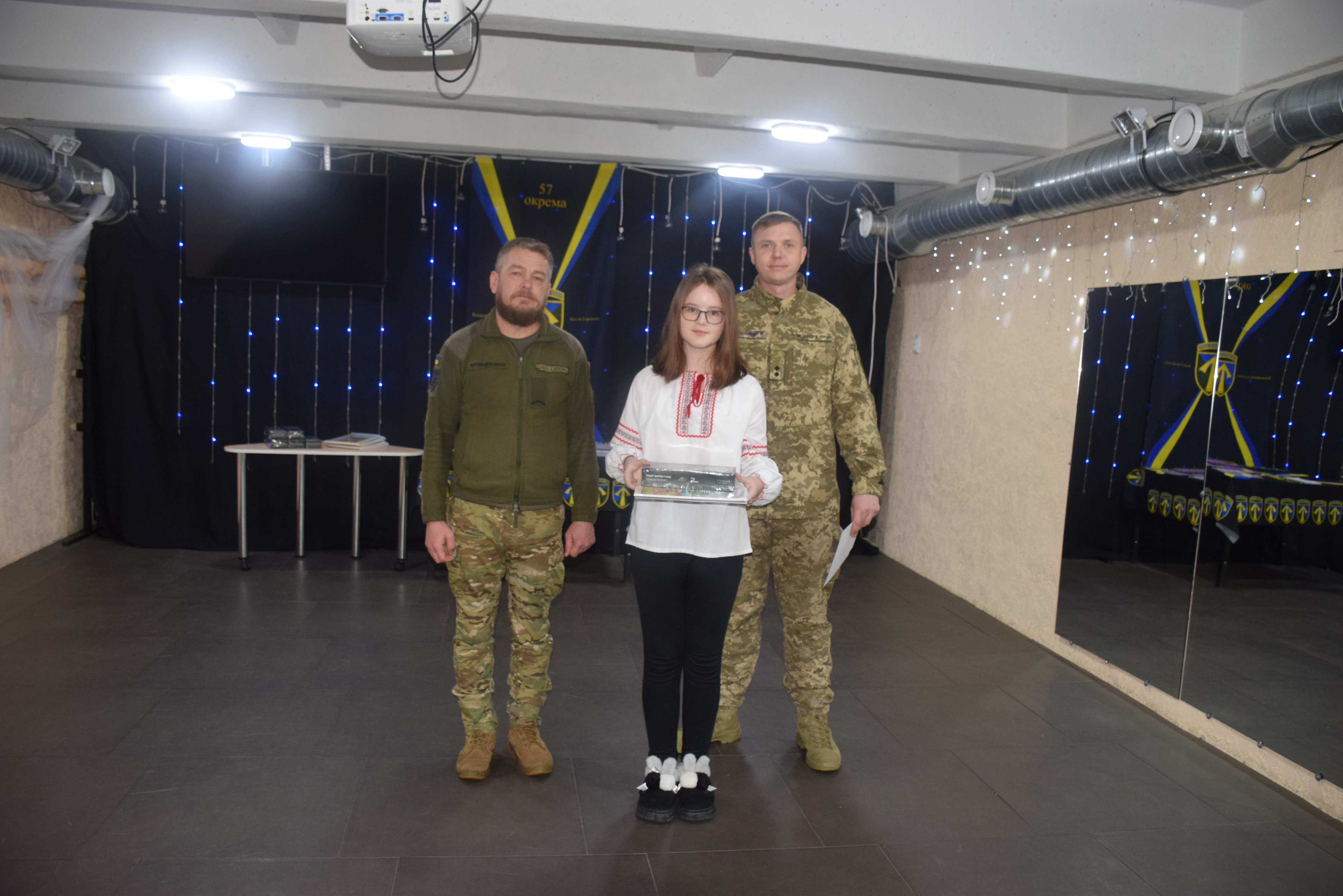 Дівчина в білому вишитому українському одязі тримає нагородження у руках, поряд стоять двоє чоловіків у військовій формі.