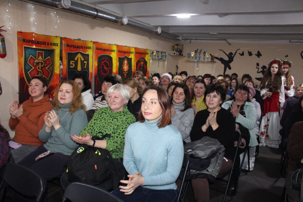 Жінки сидять у залі і аплодують виступові дівчини у національному вбранні, розташованої праворуч біля стіни.
