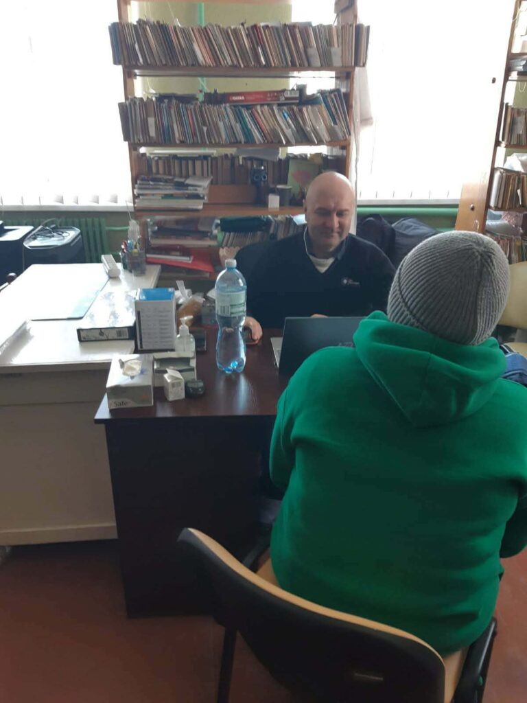 Чоловік у чорній футболці сидить за столом і працює на ноутбуку, а людина у зеленій худі і сірій шапці сидить спиною до камери.