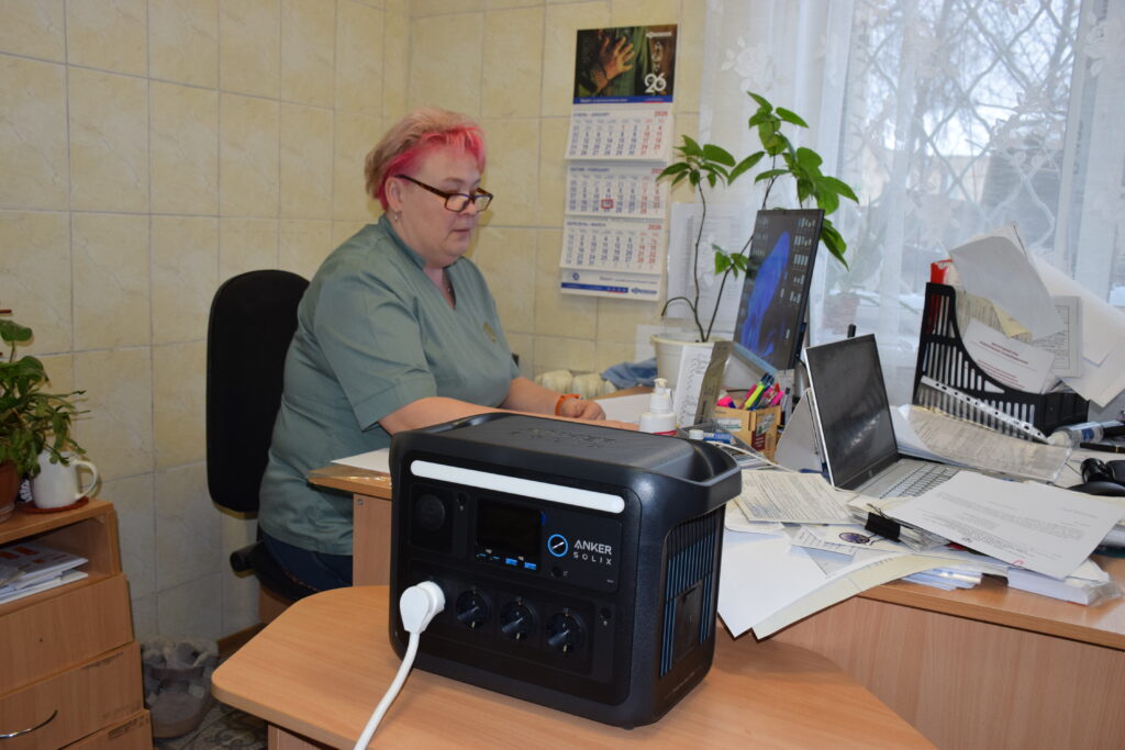 Жінка з червоним волоссям і окулярами працює за столом, навпроти неї портативна електростанція Anker Solix.