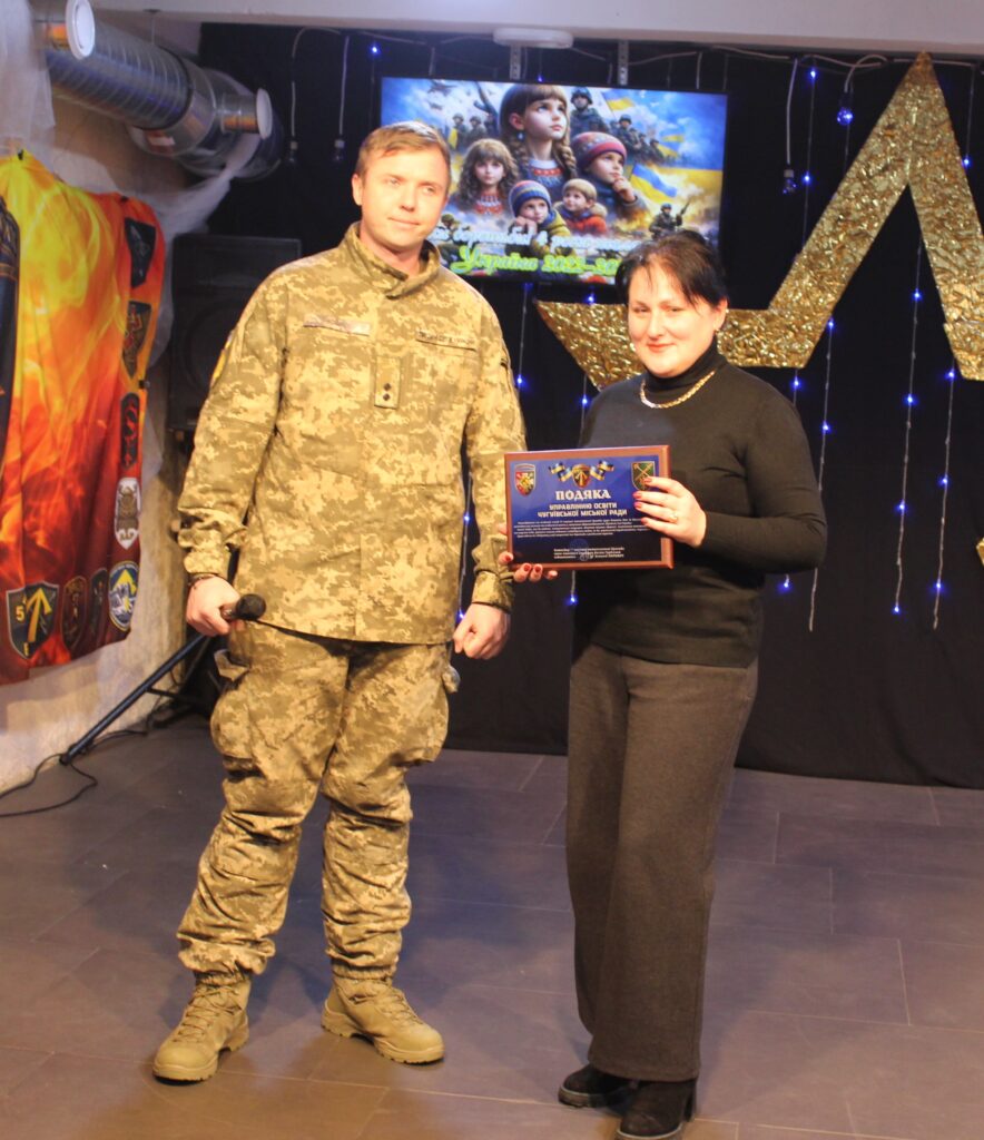 Чоловік у військовій формі та жінка у чорному одязі стоять на сцені, жінка тримає нагородну грамоту.