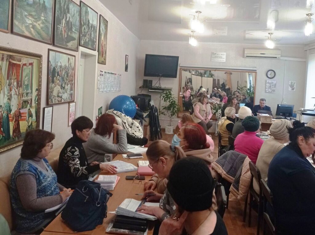 Кімната заповнена людьми, які сидять за столами та працюють із документами.