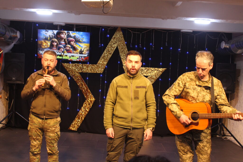 Чоловіки у військовій формі виконують музичний твір: один грає на сопілці, інший співає, третій грає на гітарі на тлі великого золотого п'ятикутника із блакитними вогниками.
