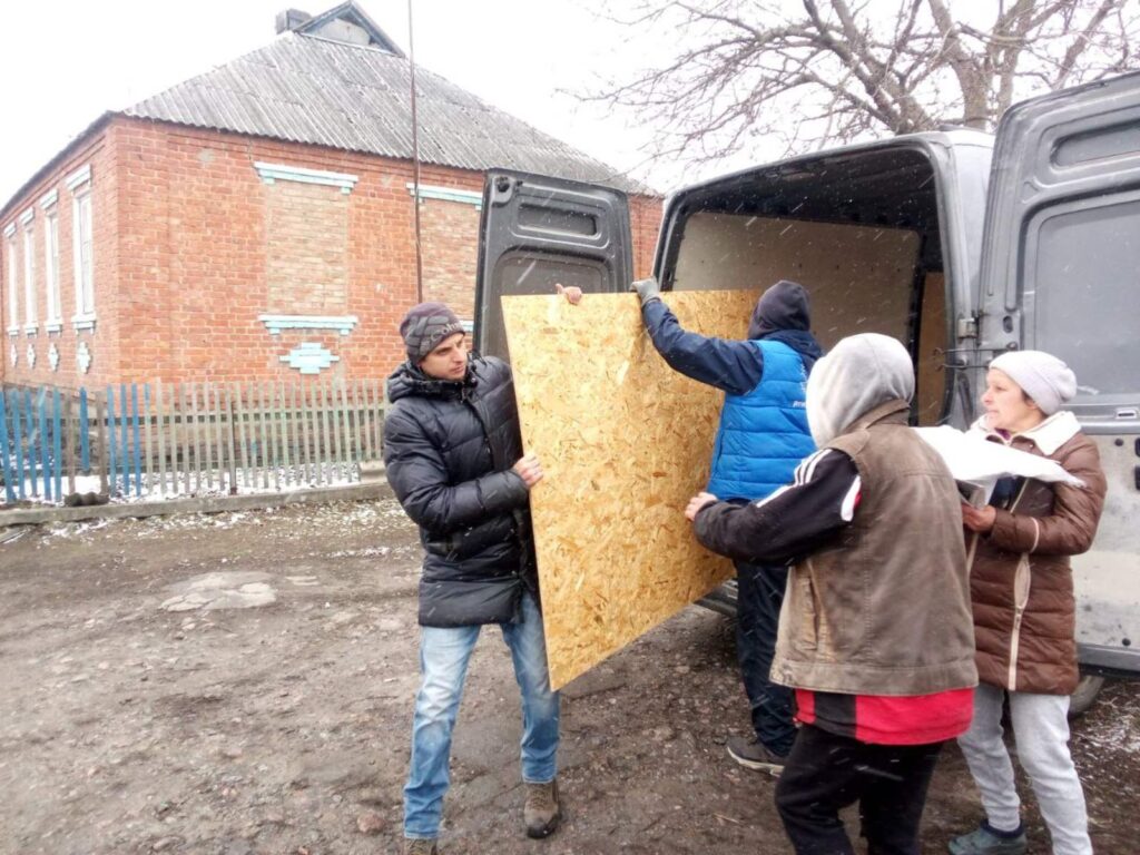 Чоловік у чорній куртці та шапці тримає велику фанеру разом із чоловіком у синій куртці та рукавицях, який завантажує її у вантажне авто.