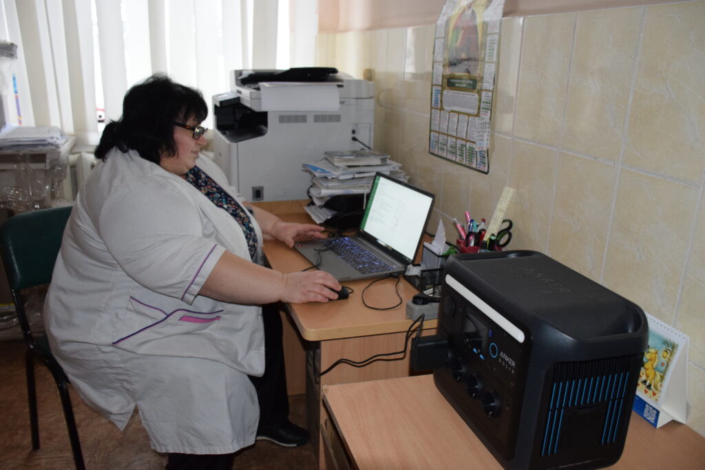 Жінка в білому халаті працює за столом з ноутбуком, сканером та офісними приладами.