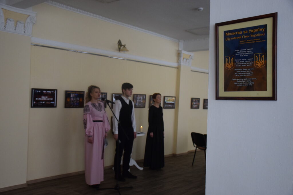 Жінка у рожевому вишиваному вбранні співає перед мікрофоном разом із чоловіком у білому сорочці та чорному жилети й жінкою у чорному вбранні.