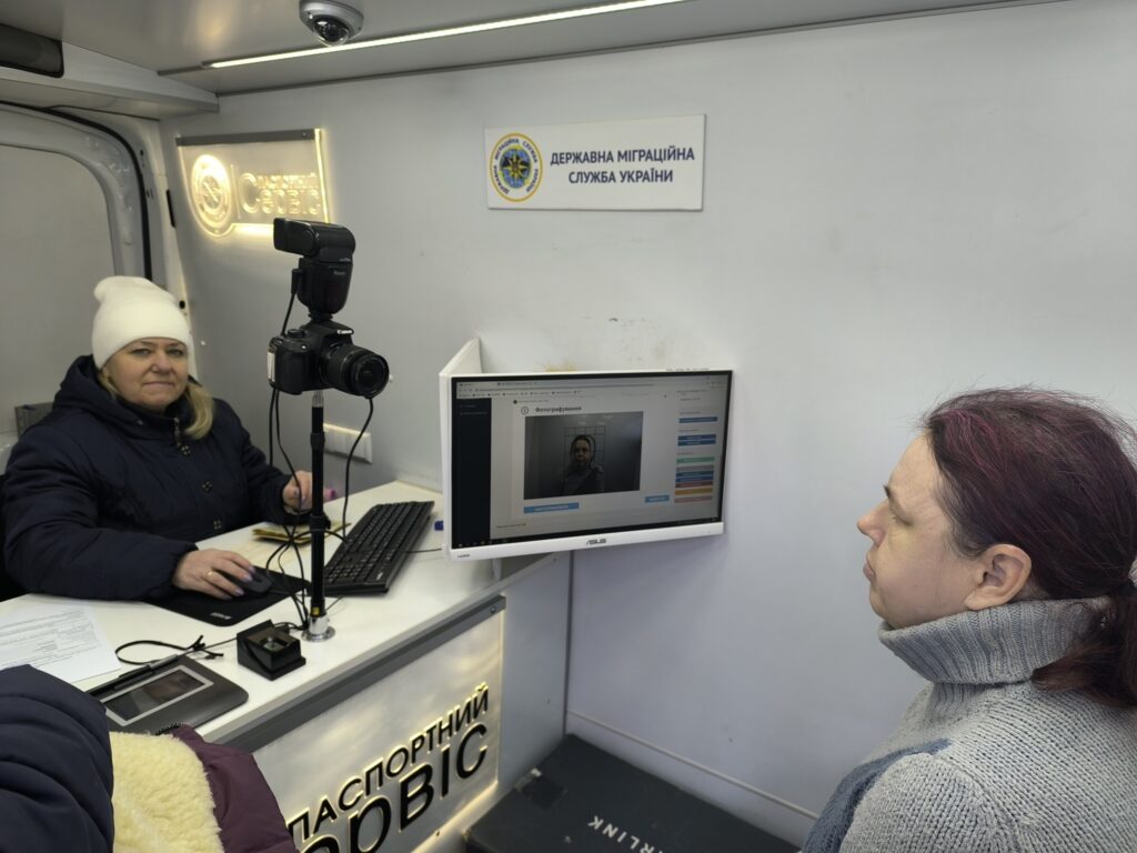 Жінка сидить біля столу та працює на комп'ютері, а інша жінка стоїть навпроти неї та дивиться на монітор із відкритим інтерфейсом для фотографування.