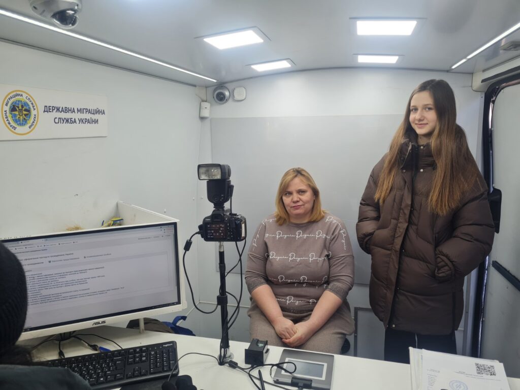 Жінка сидить біля столу з монітором та камерою, а дівчина стоїть поруч у приміщенні Державної міграційної служби України.
