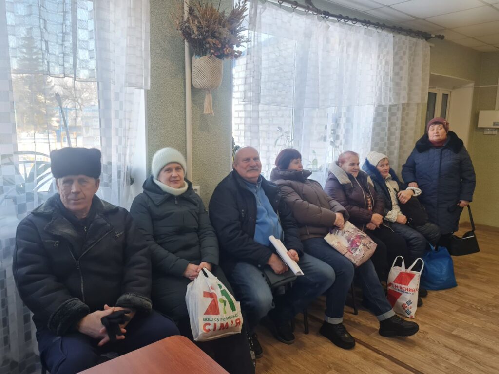 Літні люди сидять біля вікна із задертими шторами, деякі з них тримають пакети.