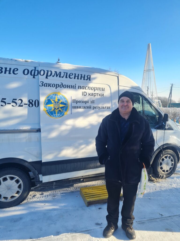 Чоловік у чорному одязі стоїть біля білого фургона з українським текстом та логотипом, що рекламує закордонні паспорти та ID-картки.