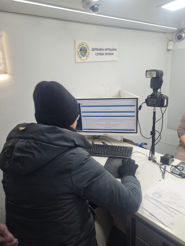 Людина в чорній шапці та куртці працює за комп'ютером у приміщенні Державної міграційної служби України.