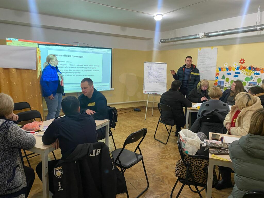 Жінка біля проектора та чоловік біля білої дошки ведуть семінар для групи людей, сидячих за столами.