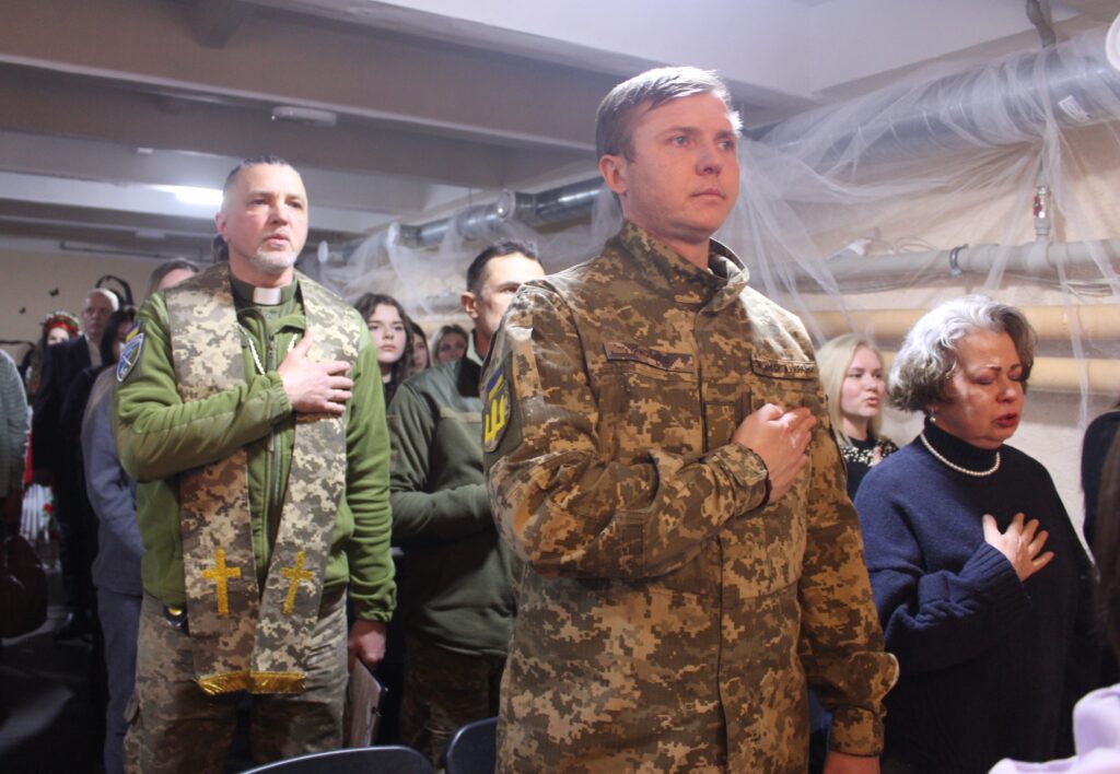 Чоловік у військовій формі стоїть із рукою на серці серед інших людей у приміщенні.