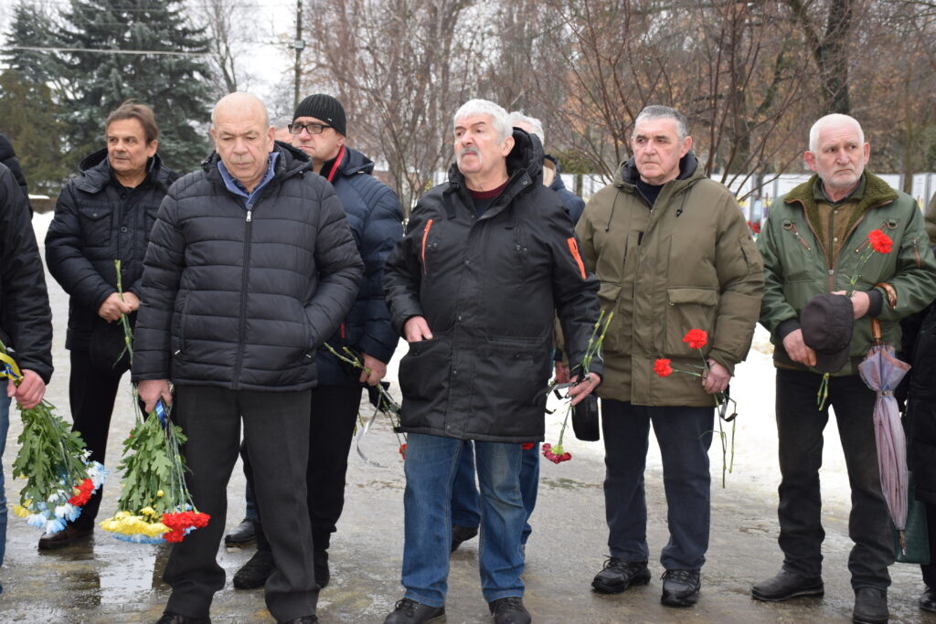 Чоловіки похилого віку стоять у колону і тримають квіти у руках під час проведення урочистої церемонії покладання квітів.