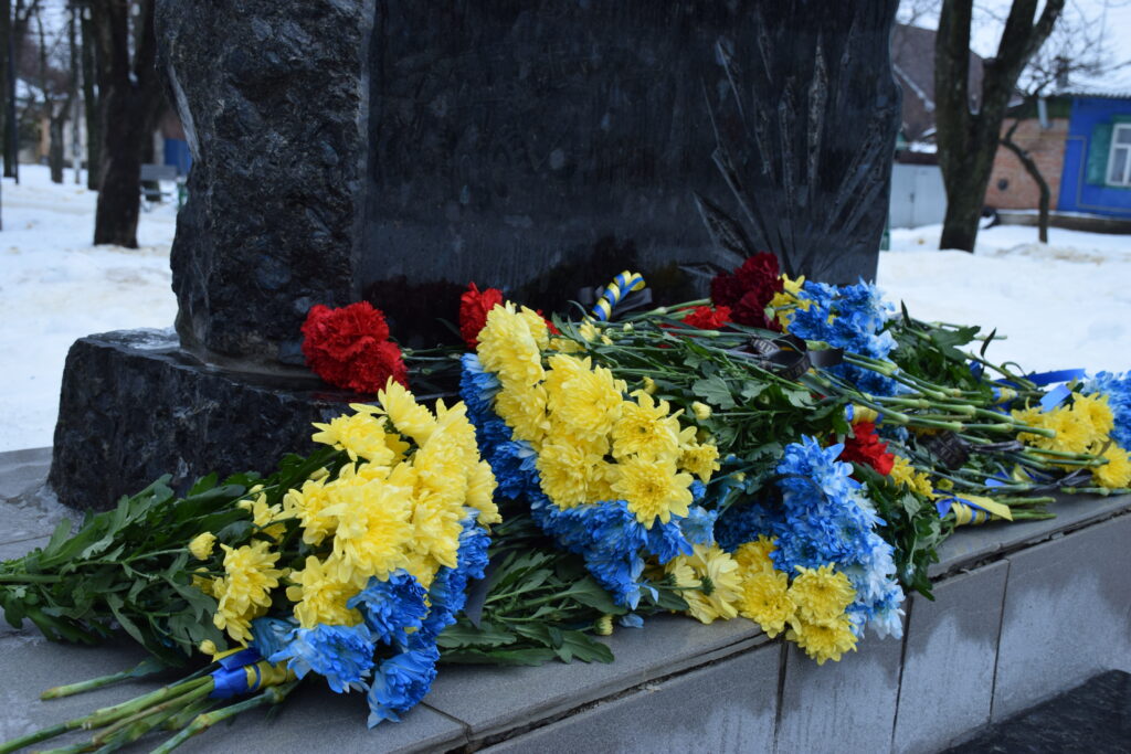 Букет із жовтих, синіх і червоних квітів біля підніжжя чорного пам'ятника на тлі зимового пейзажу.