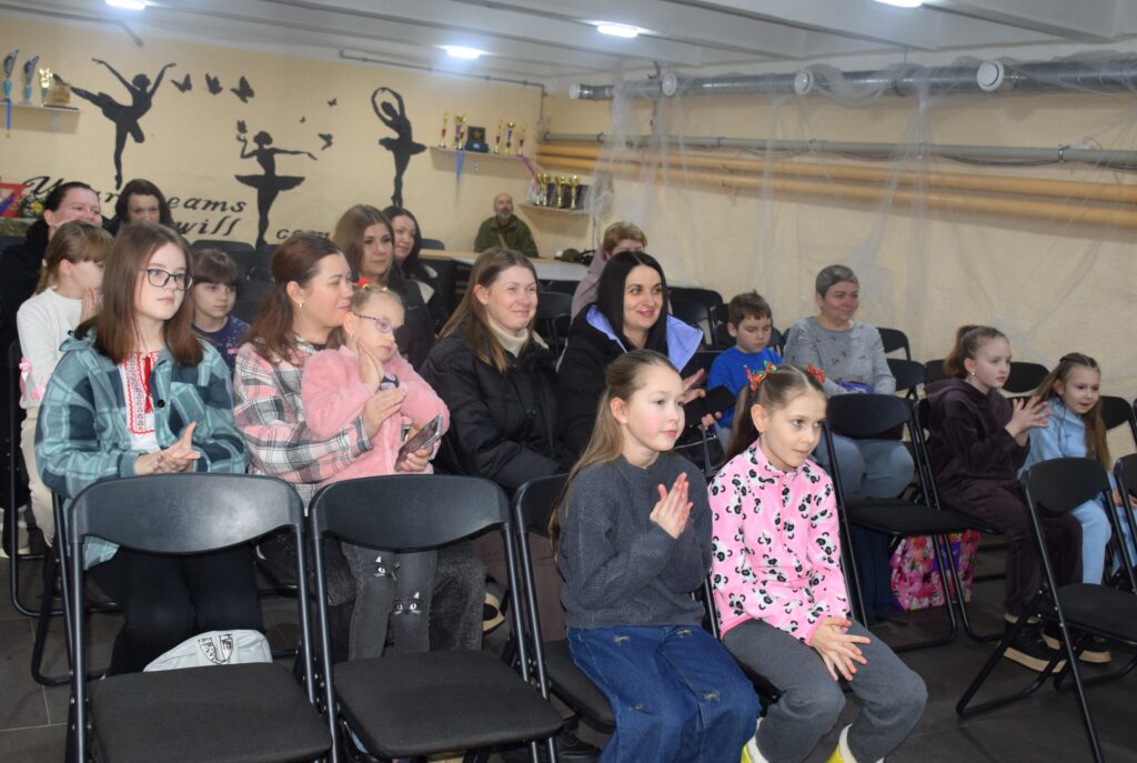 Жінки та діти сидять у чорних кріслах, деякі з яких аплодують, у кімнаті з кремовою стіною та чорними силуетами балерин.