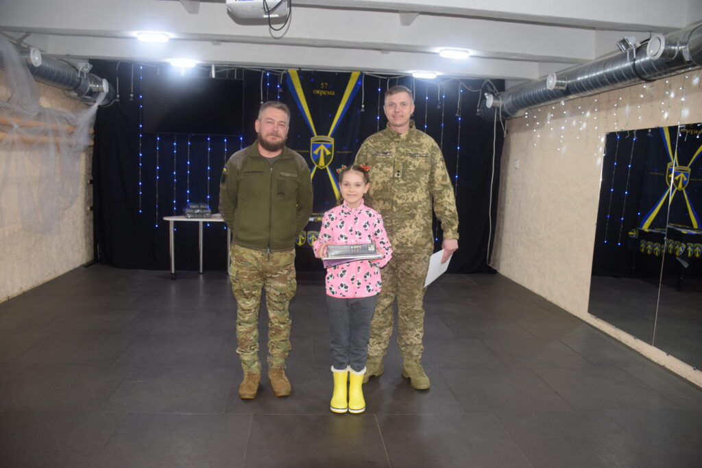 Двоє чоловіків у військовій формі стоять по обидва боки від дівчинки, яка тримає в руках радіостанцію.