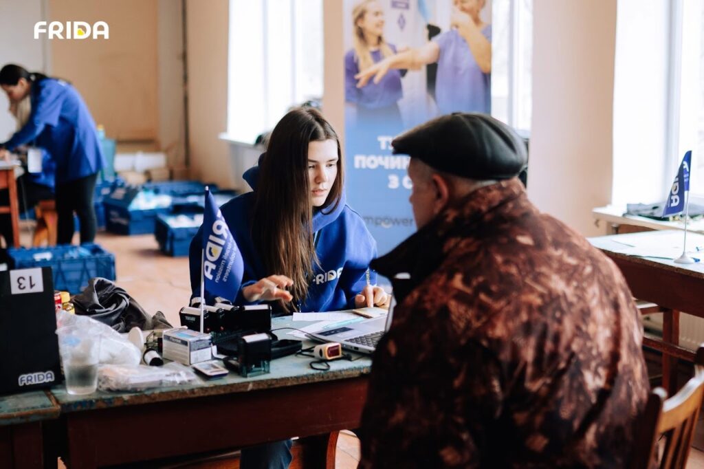 Дівчина у синьому худі працює з чоловіком за столом, серед різних предметів, у приміщенні з плакатом та іншими людьми на задньому плані.
