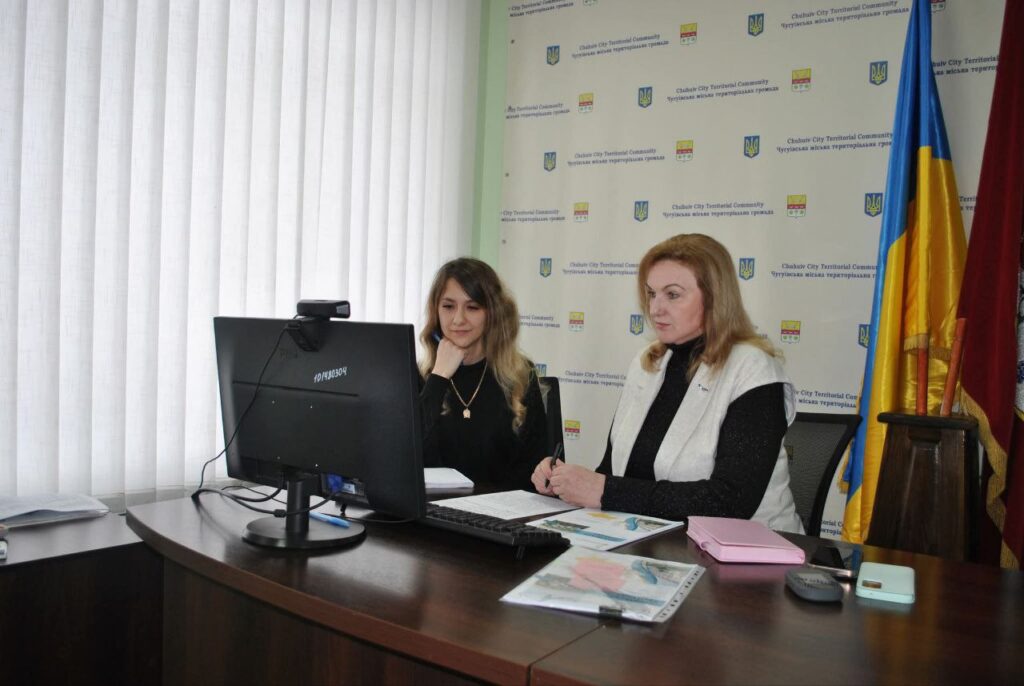 Дві жінки сидять за столом із комп'ютером, розглядаючи документи.