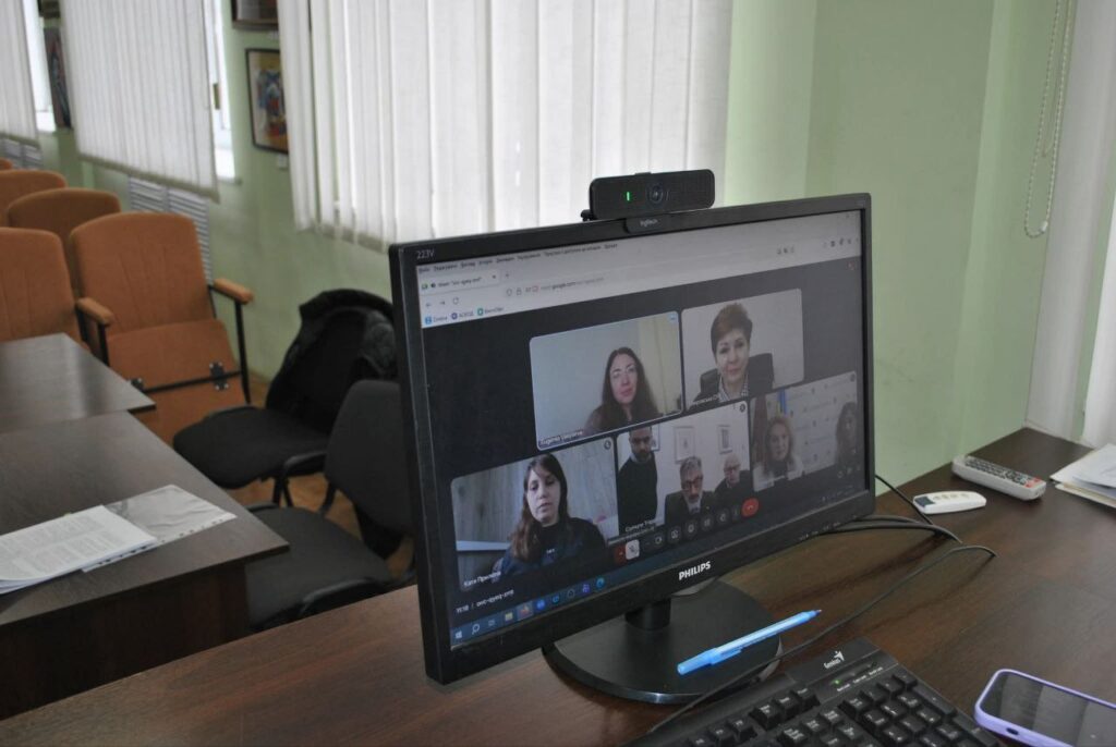 Монітор із вбудованою вебкамерою на столі відображає відеоконференцію за участю чотирьох жінок та трьох чоловіків.