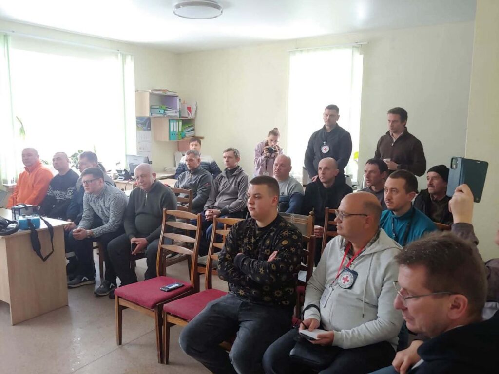 Чоловіки різних вікових груп сидять і стоять у приміщенні, деякі тримають телефони, а один робить знімок.