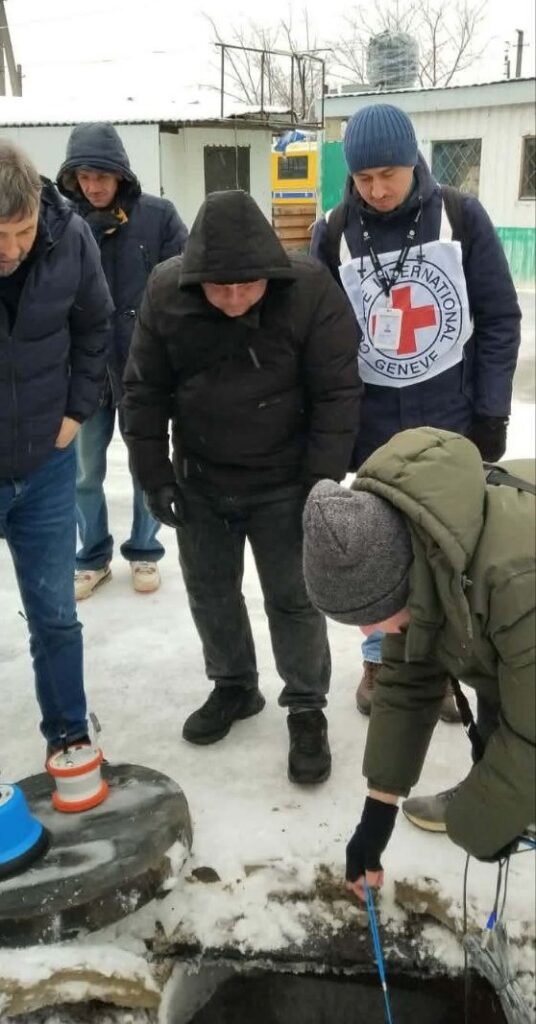 Чоловік у зеленому одязі вимірює глибину криниці за допомогою синього шнура, а троє чоловіків у темному одязі та жінка з мішком Червоного Хреста уважно спостерігають.