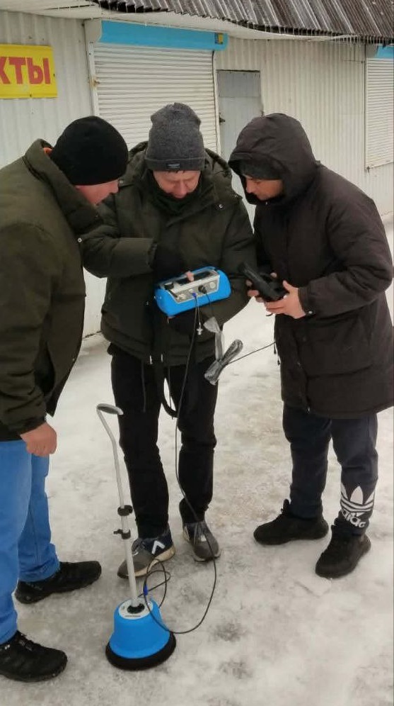 Три чоловіки в зимовому одязі працюють із георадарним обладнанням на снігу біля будівлі.