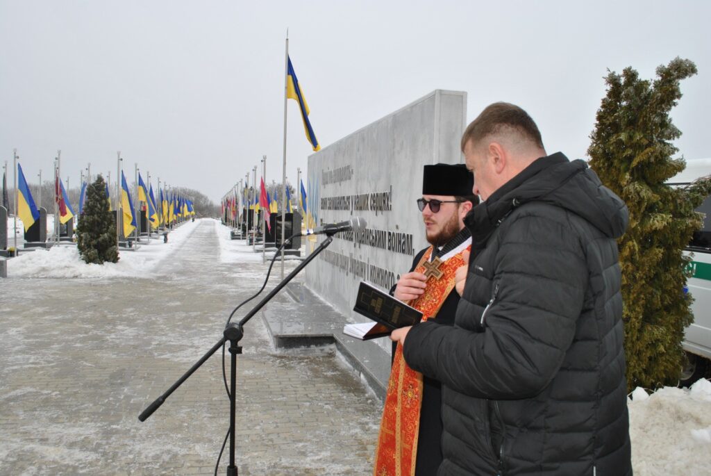 Чоловік у чорній куртці читає книгу, мікрофон розташований перед ним; позаду нього інший чоловік у чорному вбранні та жовтогарячій рясі.