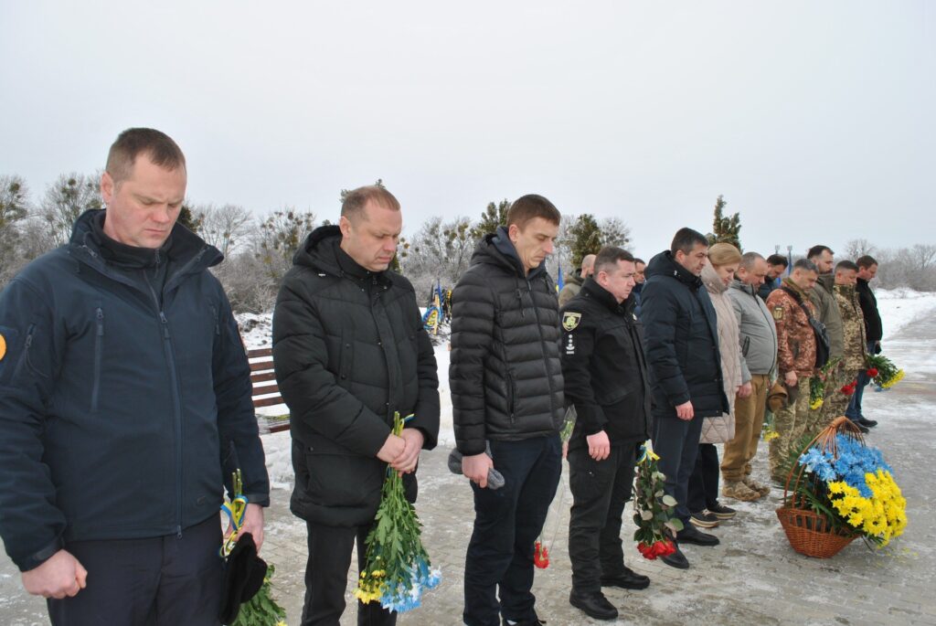 Кілька людей стоять у рядку, опустивши голови і тримаючи квіти, ймовірно під час меморіальної церемонії.