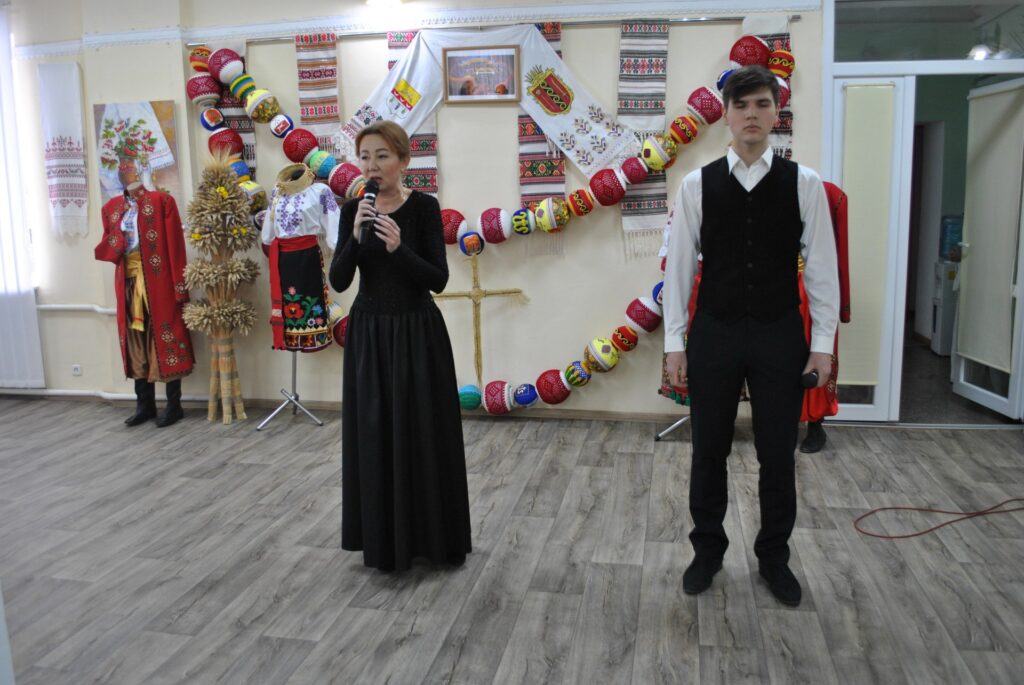 Жінка у чорній сукні тримає мікрофон перед мікрофоном, а чоловік у чорному піджаку стоїть поруч.