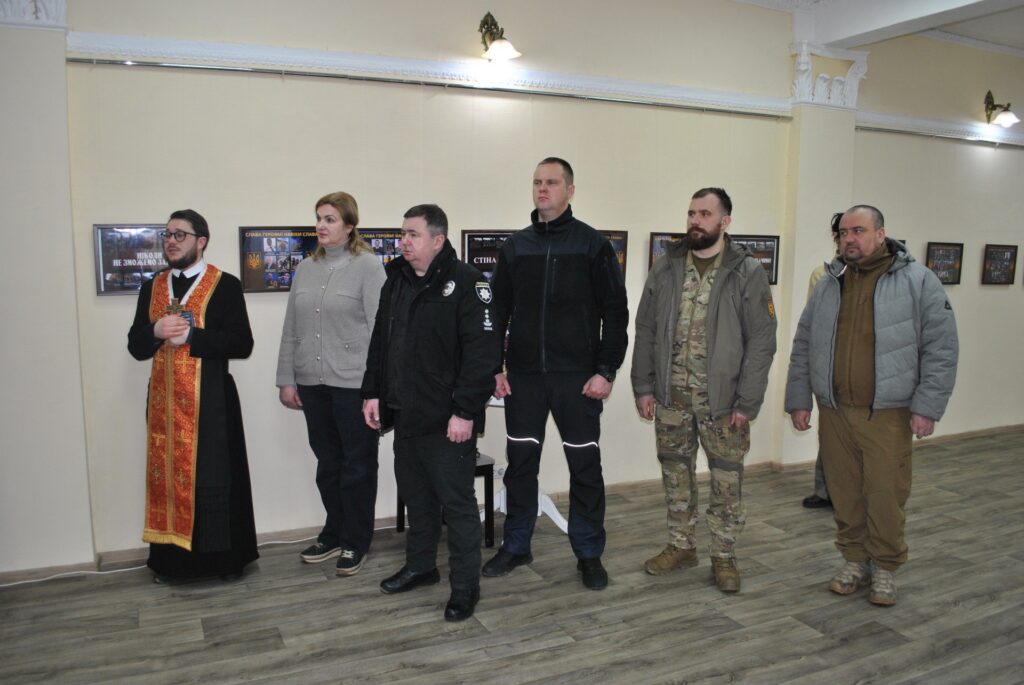 Шістьох людей різного віку та в різному одязі стоять біля стіни із світлими картинами.