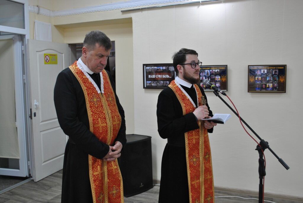 Чоловік у окулярах виступає перед мікрофоном із книгою в руках, поряд інший чоловік із опущеними вниз очима.