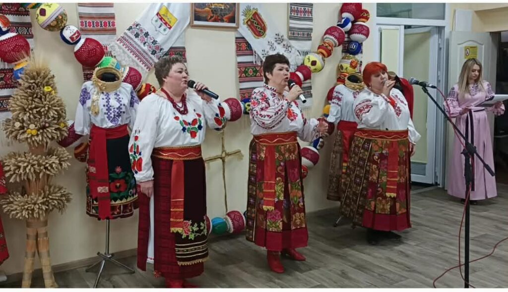 Жінки у вишитих українських національних костюмах співають біля мікрофонів, прикрашених декором із вишитими сорочками та іншими елементами народного вбрання.
