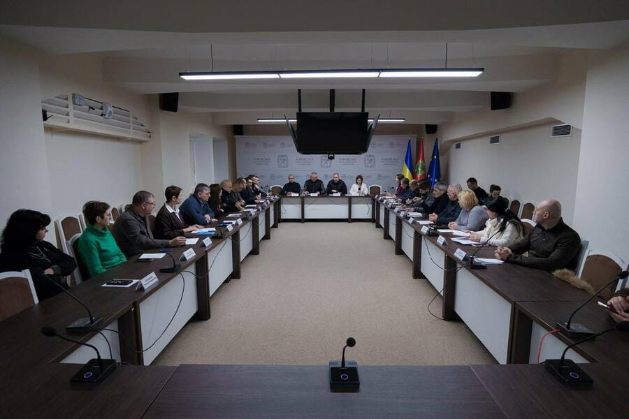 Багато людей сидять за довгими столами у формі літери П у приміщенні із прапорами Євросоюзу та України.
